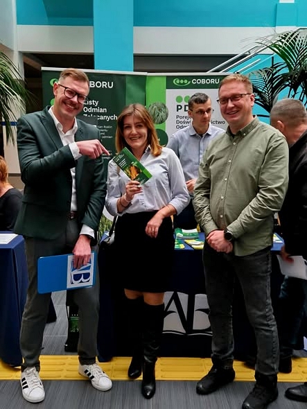 Prelegent tiktoker Rolnik na każdą okazję wskazujący palcem na listę odmian zalecanych którą trzyma pracownica SDOO Cicicbór na tle stoiska stacji